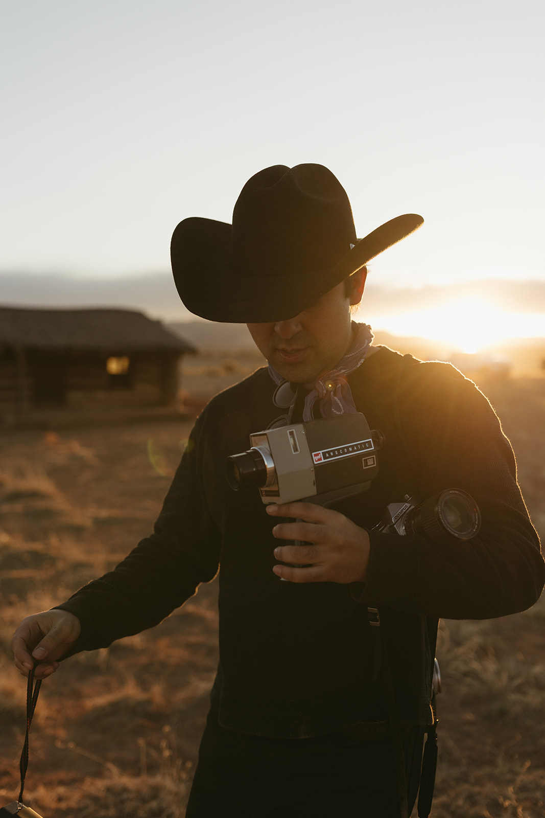 Frankie Gomez in a black cowboy hat holding a vintage Super 8 camera at golden hour, with an old adobe cabin and the New Mexico sun behind him